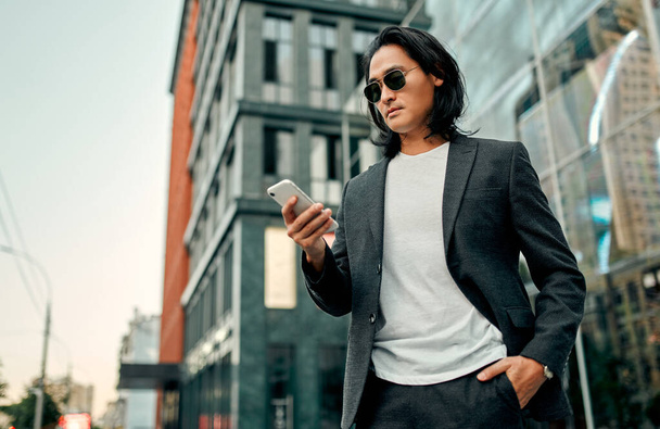 Asian businessman in the city. Confident young man standing outdoor in suit and sunglasses with smart phone in hands - Photo, Image