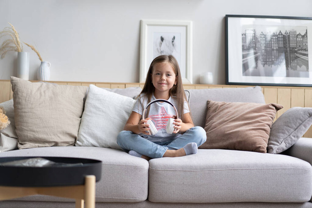Adorable small schoolgirl sitting on sofa, wearing modern wireless headphones, listening to favorite music - Foto, Bild