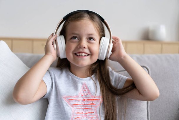 Adorable small schoolgirl sitting on sofa, wearing modern wireless headphones, listening to favorite music - Fotografie, Obrázek