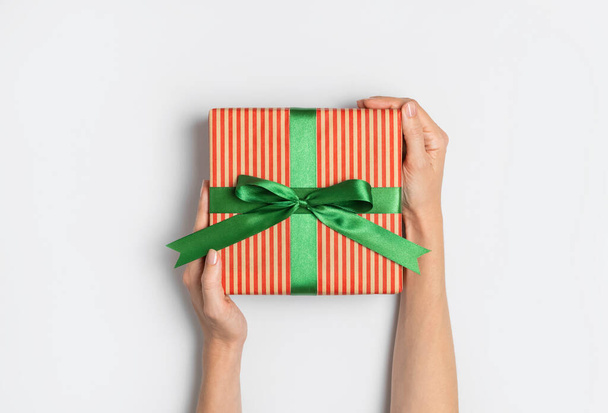 Female hands hold gift box in striped craft paper with green bow on light background flat lay top view copy space. Holiday present, christmas, new year, birthday, valentine's day, 8 march. Surprise. - Fotó, kép