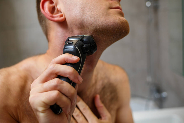 男は鏡の前で電動カミソリで顔を剃る。皮膚刺激。風呂の手順 - 写真・画像