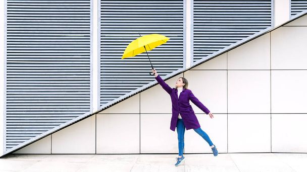 Beyaz geometrik arka planda sarı şemsiyeyle atlayan mor ceketli sarışın bir kadın. Metnin için alanı kopyala. Kış konsepti. - Fotoğraf, Görsel