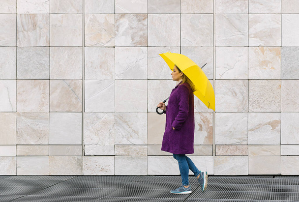 Blonde woman wearing a purple coat walking with a yellow umbrella on a stone architecture background. Copy space for your text. Winter concept. - Foto, immagini