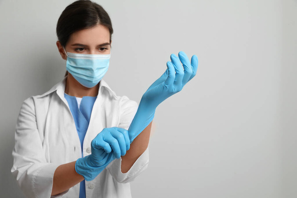 Doctor in protective mask putting on medical gloves against light grey background. Space for text - Фото, зображення