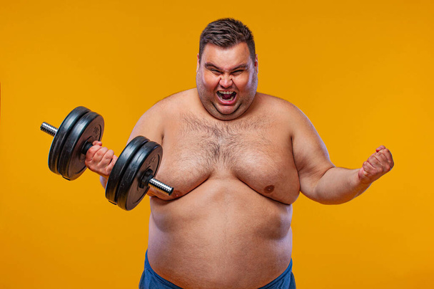 Fat man doing workout traning with dumbbells. Happy guy isolated over yellow background. - Photo, image