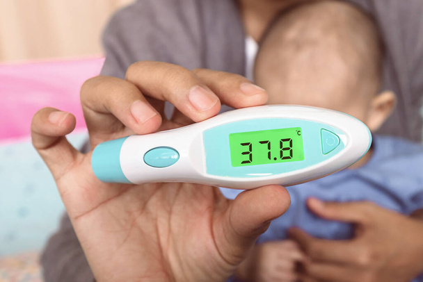 Asiatische Mutter hält ein digitales Thermometer Messung ihres Sohnes, Fokus auf das Thermometer - Foto, Bild