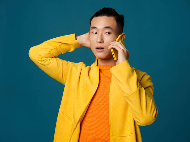 Asian man holds his hand behind his head and talks on the phone on a blue background - Fotó, kép