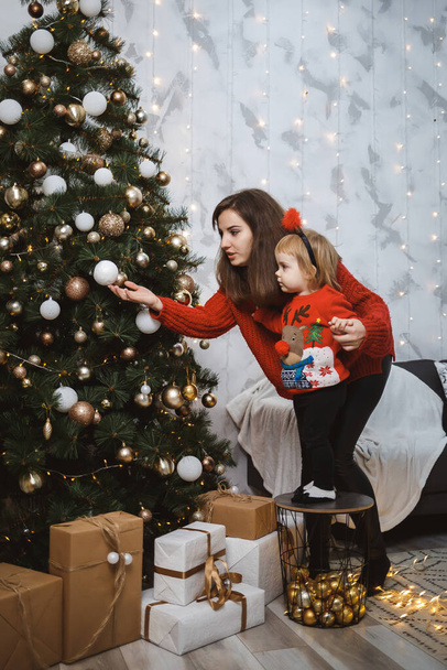 Anya és lánya piros meleg pulóverben díszítik a karácsonyfát. Boldog anyaságot! Meleg családi kapcsolatok. Karácsonyi és szilveszteri lakosztály. Szerelem. Családi fogalom. - Fotó, kép
