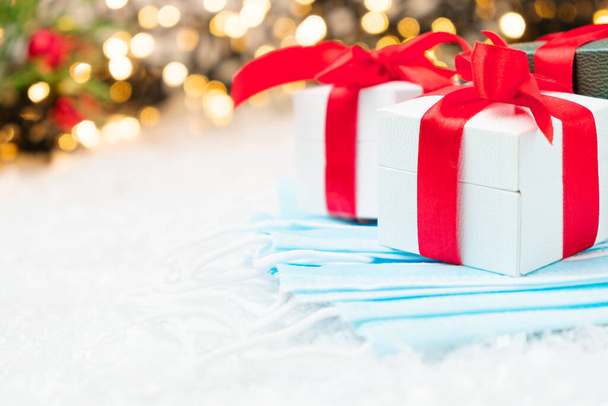 Regalos o caja de regalo con una cinta roja y máscaras azules contra el coronavirus en la nieve con bokeh dorado de luces, cono y árbol de Navidad en el fondo. Ambiente de año nuevo. Copiar espacio. - Foto, imagen