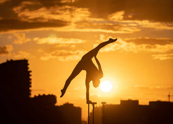 Flexible acrobat doing handstand on the cityscape background during dramatic sunset. Concept of willpower, control and dream - 写真・画像