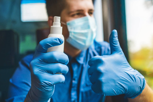 A man on the background in a protective mask holds an antiseptic in his hand in medical gloves and shows a cool thumb up - Fotografie, Obrázek