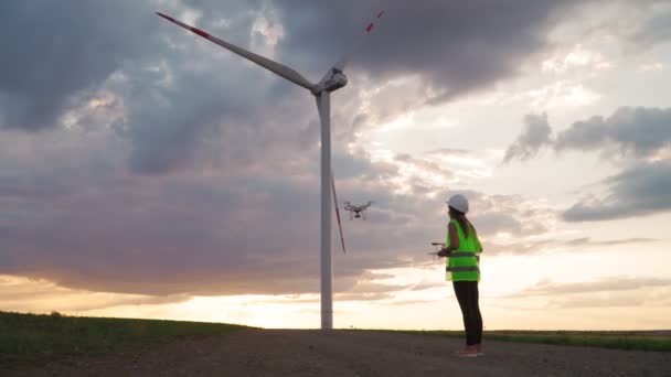 Femme Ingénieur écologie en uniforme et casque tenant joystick contrôlant drone volant travaillant au moulin à vent sur beau fond de coucher de soleil. Alternative à l'énergie électrique. - Séquence, vidéo