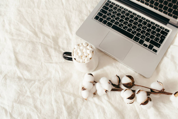 Laptop and cup of cocoa with marshmallows, cotton flowers on white bed. Work at home concept. Autumn, fall, winter composition. Flat lay, top view, copy space. - Foto, Imagem