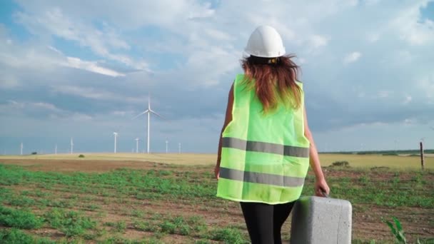 手に特別な機器を備えた制服やヘルメットの女性プロの生態エンジニアは、美しい空とフィールドの背景に風車を提供するために行きます。代替エネルギーの概念。スローモーション. - 映像、動画