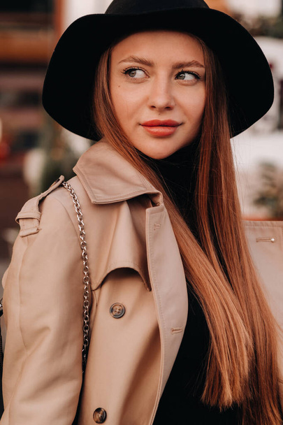 Portrait of a girl in a beige coat and black hat on a city street. Women's street fashion. Autumn clothing.Urban style. - Photo, image