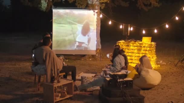 Przyjaciele spędzający razem wieczór filmowy oglądając filmy na ekranie kina na świeżym powietrzu wieczorem jedząc pizzę i pijąc piwo - Materiał filmowy, wideo