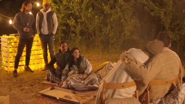 Volledig shot van twee gelukkige koppels en hun vrienden die het weekend buiten praten, bier drinken en ontspannen onder de open lucht bedekt met dekens 's nachts - Video