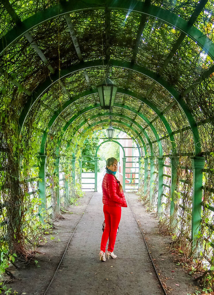Veselá mladá žena v červeném kráčí po městském parku. Dívá se na fotografa s úsměvem. - Fotografie, Obrázek
