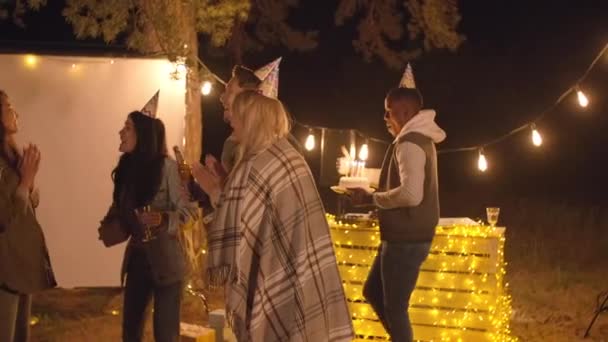 Grupa wesołych młodych ludzi w kapeluszach imprezowych śpiewa piosenkę urodzinową na świeżym powietrzu, podczas gdy ich afrykańscy przyjaciele przynoszą tort urodzinowy z płonącymi świecami w letni wieczór - Materiał filmowy, wideo