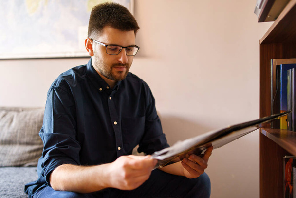 Adult caucasian man sitting on the sofa or bed at home wearing blue shirt and eyeglasses reading book at home study in day front view low angle - Photo, Image
