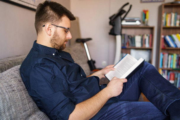 Adult caucasian man sitting on the sofa or bed at home wearing blue shirt and eyeglasses reading book at home study in day side view - 写真・画像