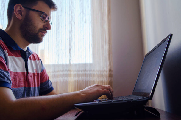 Hombre joven caucásico adulto con camiseta y anteojos sentados junto a la mesa pelo corto y barba trabajando en computadora portátil en casa navegando por internet vista lateral - Foto, Imagen