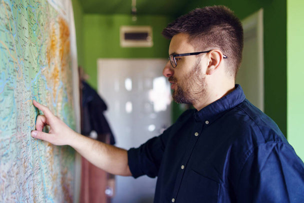Портрет молодого кавказца мужчины, стоящего рядом с большой картой, висящей дома на стене, указывающей пальцем на место преступления в рубашке и очках в дневном боковом виде - Фото, изображение