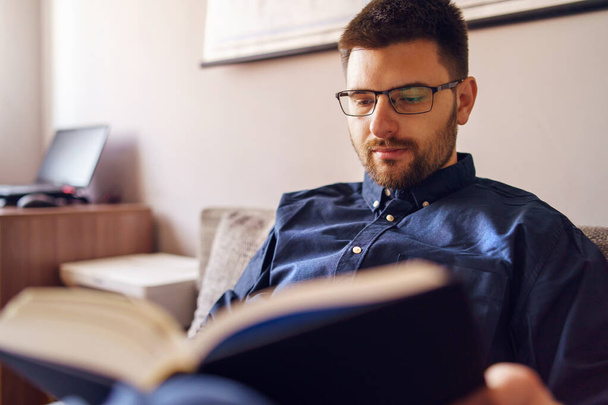 Um homem caucasiano adulto sentado no sofá-cama em casa lendo o estudo do livro aprendendo sozinho durante o bloqueio de quarentena em um dia vestindo camisa e óculos e vista frontal da barba - Foto, Imagem
