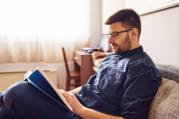 Egy felnőtt kaukázusi férfi otthon ül a kanapén, és a könyvet olvassa. Egyedül tanul a karantén alatt, egy nap ingben, szemüvegben és szakállas oldalnézetben. - Fotó, kép