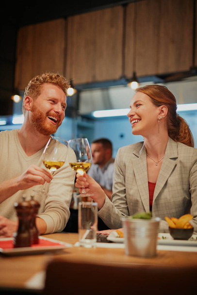 Νεαρό χαμογελαστό, ευτυχισμένο ζευγάρι ερωτευμένο να κάθεται σε ένα εστιατόριο για δείπνο και να κάνει πρόποση με λευκό κρασί. - Φωτογραφία, εικόνα