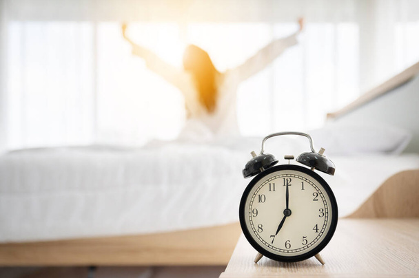 Καλημέρα νέα μέρα. ξυπνητήρι ξυπνήσει και γυναίκα κάθεται σώμα τεντώσει στο κρεβάτι δίπλα από το παράθυρο στην κρεβατοκάμαρα - Φωτογραφία, εικόνα