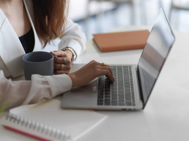 Side view of female working with laptop and holding coffee cup on office desk - Photo, image