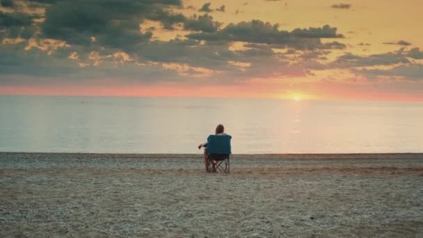 Vista trasera de la chica admirando la puesta de sol en el mar sentado en silla de turismo plegable - Metraje, vídeo