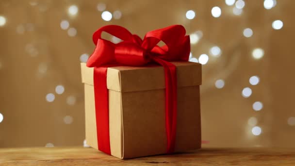Подарочная коробка, завернутая в красную ленту, стоит на коричневом фоне. Рождественская гирлянда сверкает на заднем плане - Кадры, видео