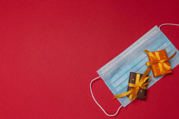 Голубая медицинская маска с двумя маленькими коробочками в цветной бумаге с горошек перевязаны желтой лентой с подарком на красном фоне. Подарок или сюрприз на Рождество, день рождения или любую годовщину.  - Фото, изображение