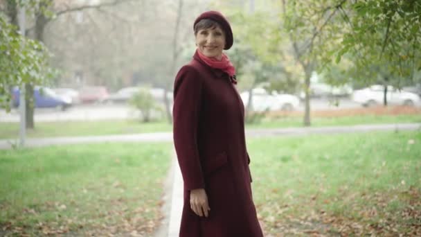 Portrait of gorgeous senior woman posing on autumn day outdoors. Happy beautiful Caucasian female retiree standing outdoors, looking at camera and smiling. Natural beauty concept. - Video