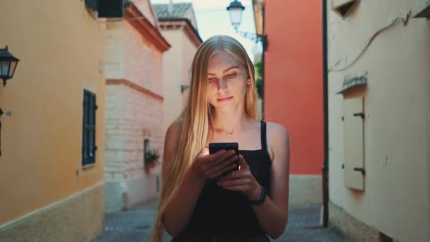 Blondýny žena čte něco na smartphone při chůzi po ulici - Záběry, video