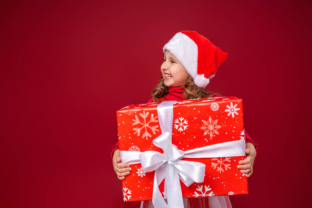 Happy little girl 4 years old in a Santa hat on a bright red background. funny child holding a gift in his hands looks away to Give gifts. New year sales, advertising, discounts. - Foto, Imagem