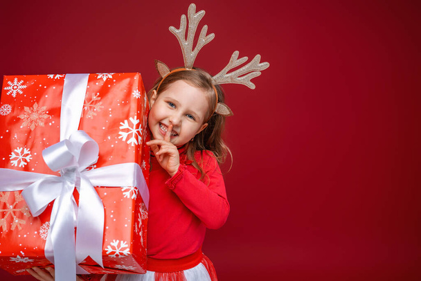Happy little girl in reindeer antlers on a red background. a cheerful child, peeking out from behind box and pressing finger to his mouth. Secret. Christmas sales, advertising, discounts. Copy space. - Foto, Imagen