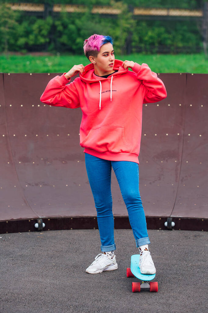 Portrait of a trendy pretty young girl dressed in pink hoodie and jeans standing next to the skateboard court with her blue plastic skateboard. - Zdjęcie, obraz