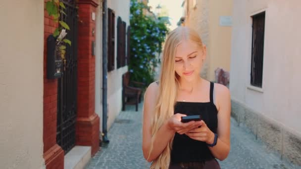 Ładna blondynka czatuje na smartfonie idąc ulicą - Materiał filmowy, wideo