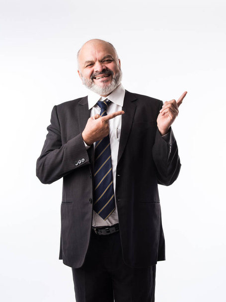 Индийский бизнесмен средних лет с бородой, одетый в элегантный черный костюм и указывающий рукой и пальцем. представление продукта, услуги или копировального пространства - Фото, изображение