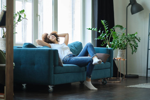 Attractive young woman relaxing on a sofa at home hands behind her head - 写真・画像