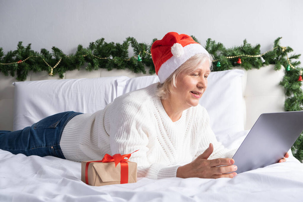Saludos en línea de Navidad. mujeres mayores felices en Santa sombrero en casa en la cama con el ordenador portátil. - Foto, imagen