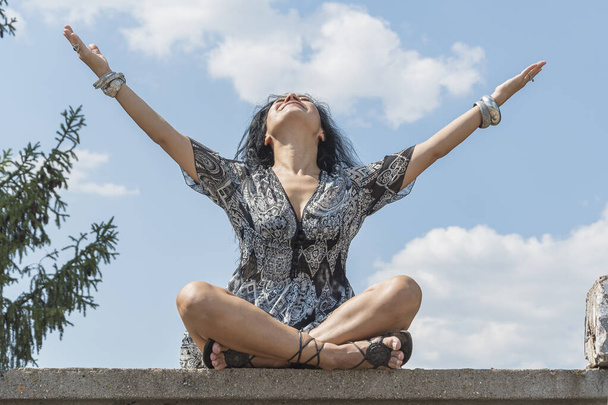 Nuori brunette tyttö istuu lootus asennossa betonipinnalla ja nostaa kätensä ja suunnata ylös taivaalle. Joogatunti. - Valokuva, kuva