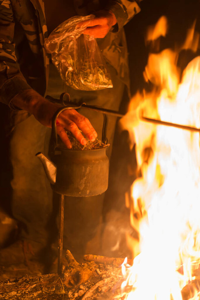 Чоловік кладе сушену траву в чайник, що кипить на яскравому вогні вночі
 - Фото, зображення