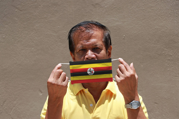 Чоловік і гігієнічна маска з зображенням прапора Уганди в руці і піднімає його, щоб закрити обличчя. Маска є дуже добрим захистом від крихітних частинок або дихальних захворювань.. - Фото, зображення