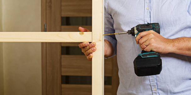 Ein Mann befestigt zwei Holzbalken mit einer selbstschneidenden Schraube. Der Tischler wickelt die Schraube mit einem Schraubenzieher. Bau eines Holzrahmens für Gipskartonverkleidungen - Foto, Bild