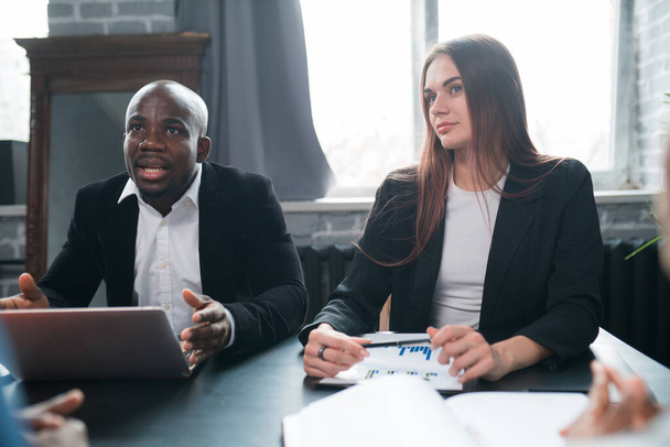 Two business partners, an African American man and a European woman are sitting at an office table at a meeting with partners. Notebook, Charts, Profit Growth Statistics - Fotografie, Obrázek
