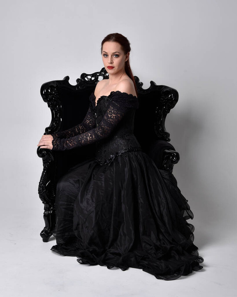 full length portrait of  woman wearing black gothic dress, sitting on a ornate black armchair. Seated pose, against a studio background. - Photo, image